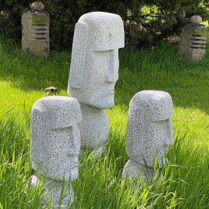 Three stone statues with stylised faces resembling Moai heads stand in tall grass, with two other sculpted stones visible in the background.